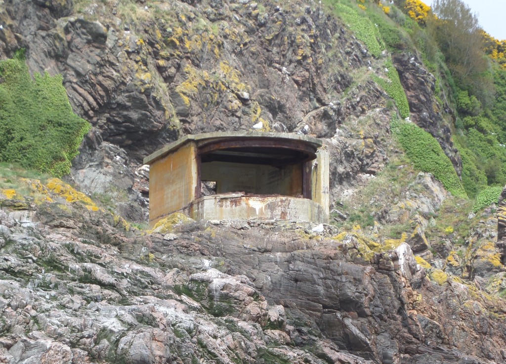 An old, rusted metal bunker nestled on a rocky cliff face, with some yellow flowers around the base and rocks.