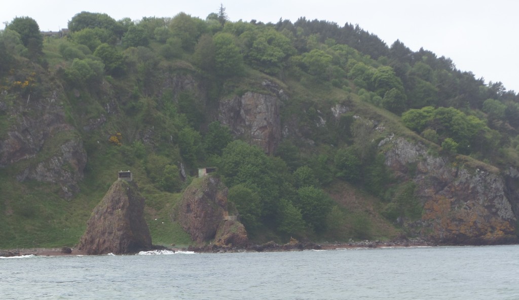 A highland coastline looking particularly grey, rocky cliffs sprouting through thick, green trees and shrubs. Two rock pillars jut up with old metal bunkers on top.