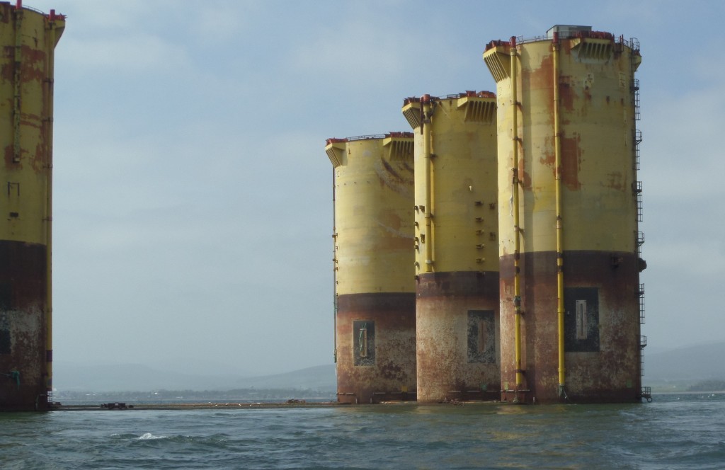 Photo of an old, decommissioned oil rig sticking up out of the water, worn and tired yellow in colour and covered with rust.
