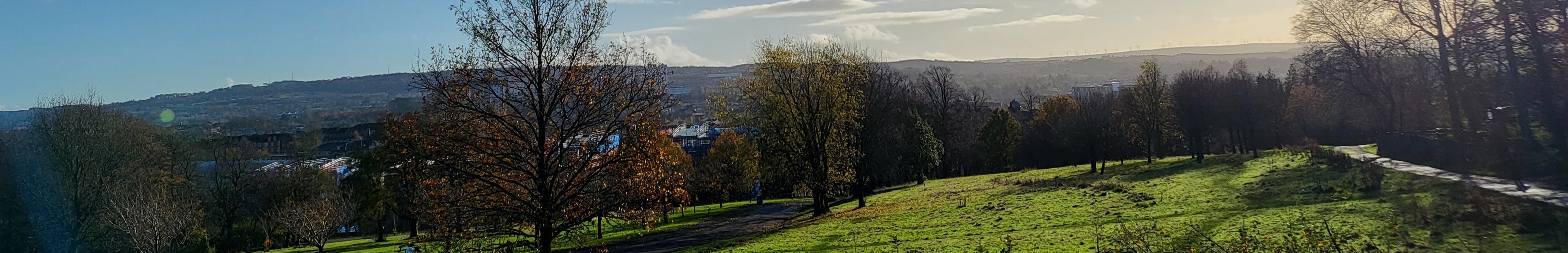 Thin banner showing a grassy field full of trees turning orange for autumn. There are mountains in the distance, a blue sky with light clouds, and a city scape just visible through the trees.